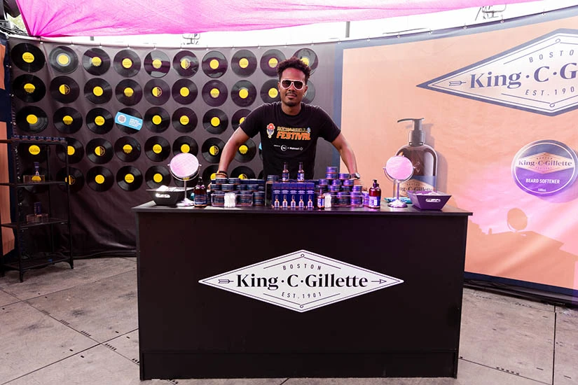 A man wearing shades and a Rock the Bells Festival t-shirt smiles as he stands behind a table that showcases the immersive King C. Gillette brand experience.