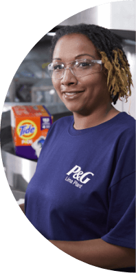A woman wearing safety glasses and a blue t-shirt that says "P&G Lima Plant" smiles with a box of Tide pods in the background.
